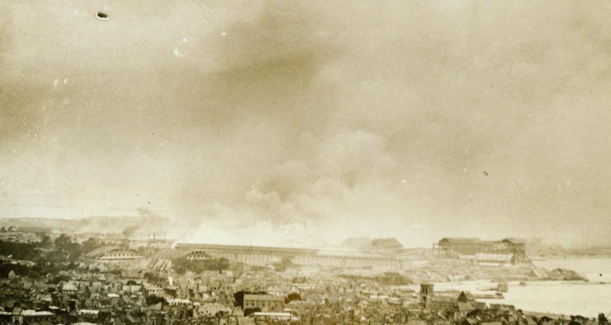 Battle Smoke Over Cherbourg, 7/1/1944. Cherbourg, France – This is a good general view of the port city of Cherbourg. Looking over the harbor as the American troops gradually closed in through the streets. Bomb-torn houses can be seen in the foreground, and smoke rises in the rear from German demolition and artillery fire. 7/1/44 (ACME Photo By Andrew Lopez, War Pool Correspondent);