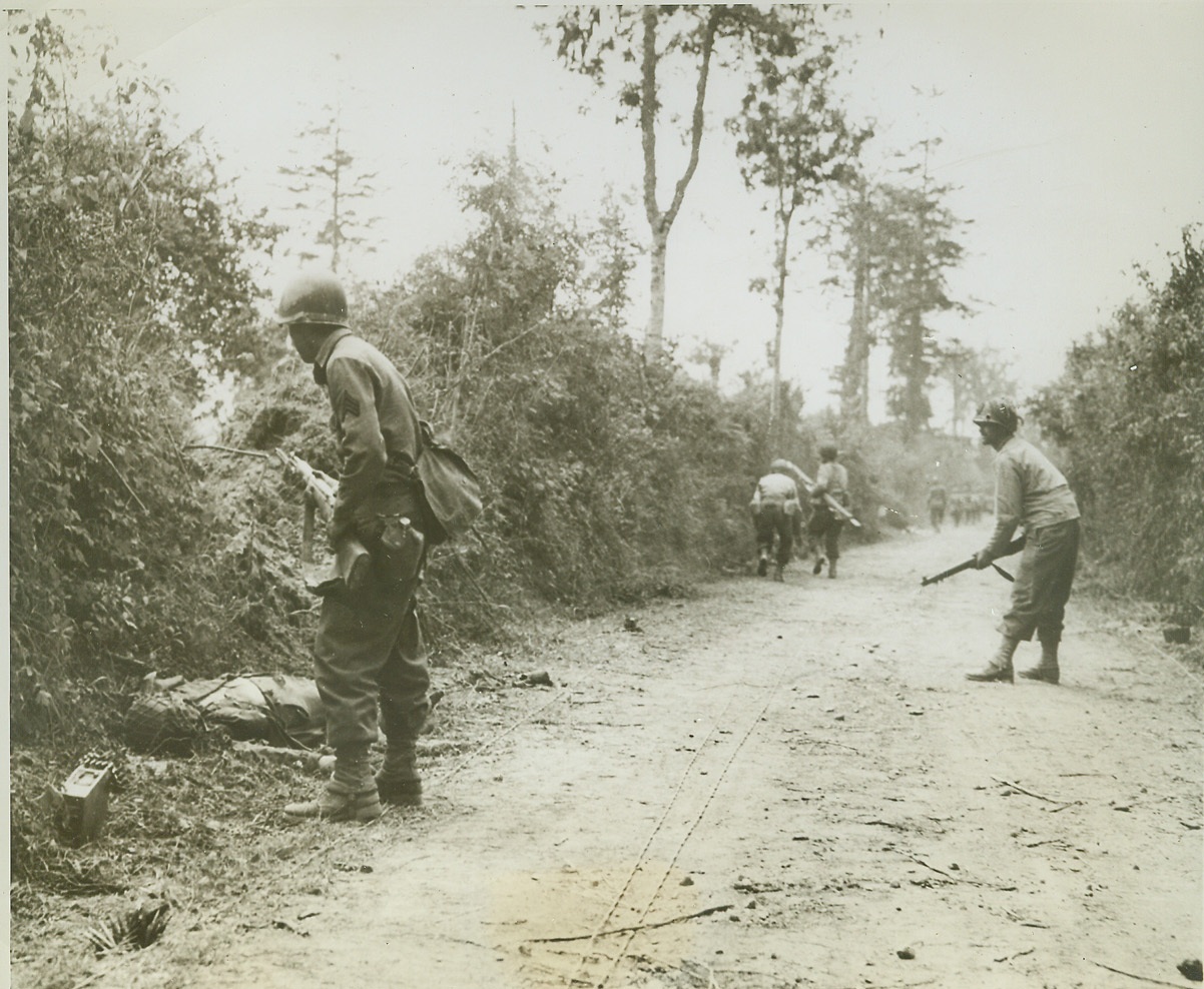 They Must Move Forward, 7/25/1944. FRANCE -- Despite the fact that one of their buddies has been killed by enemy fire, these U.S. Infantrymen continue their advance, carefully watching hedgerows for lurking snipers. Note wires put down along the road by Signal Corpsmen. Photo was taken near St. Lo. Credit: (U.S. Signal Corps Photo from ACME);