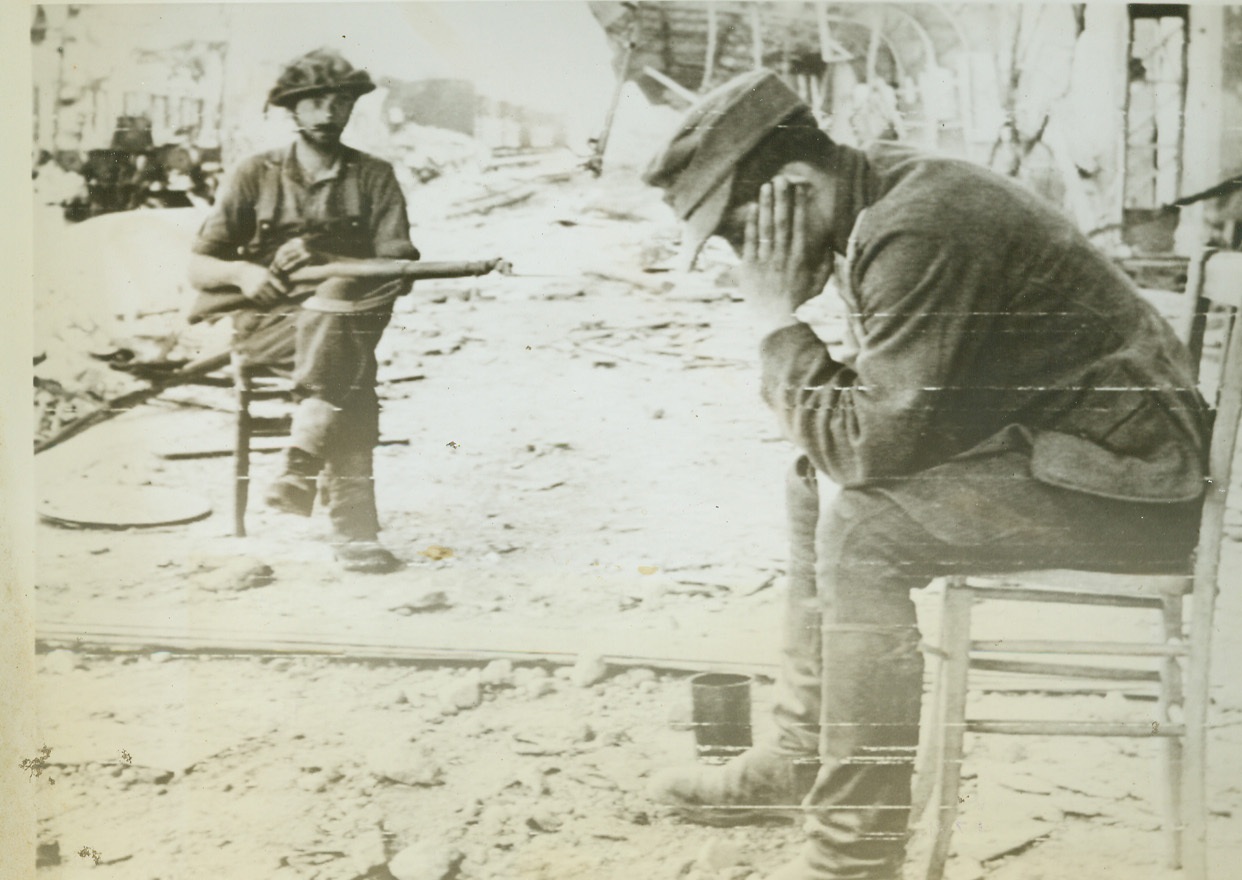 What A Cadache!, 7/23/1944. Caen, France—A weary and dejected Nazi prisoner holds his head in his hands as a Canadian soldier keeps careful watch on him. The prisoner of war cages near Caen are swollen with captives.  Credit: U.S. Army radiotelephoto from ACME;