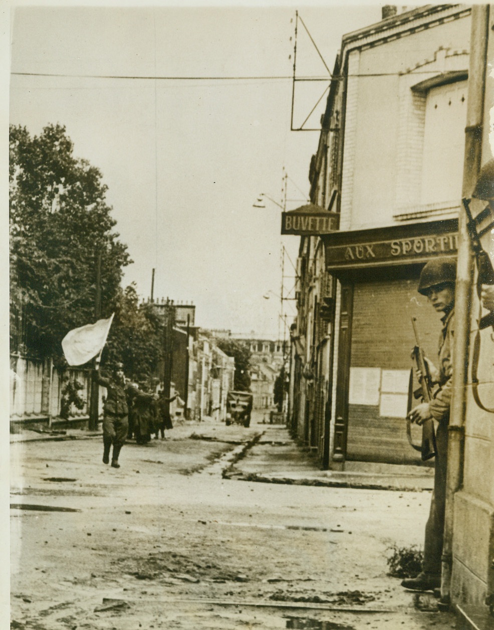 Nazis Show Their Colors, 7/1/1944. France—Defenders of a pillbox guarding a street in Cherbourg, German soldiers surrender, waving a white flag, after being knocked out of their position by Allied tank fire. Soldiers in doorway of building at right keep guns on the ready on the look out for any escape moves. Nazis carry their own wounded as they move dejectedly in defeat with their destination prison compounds.  Credit: ACME;