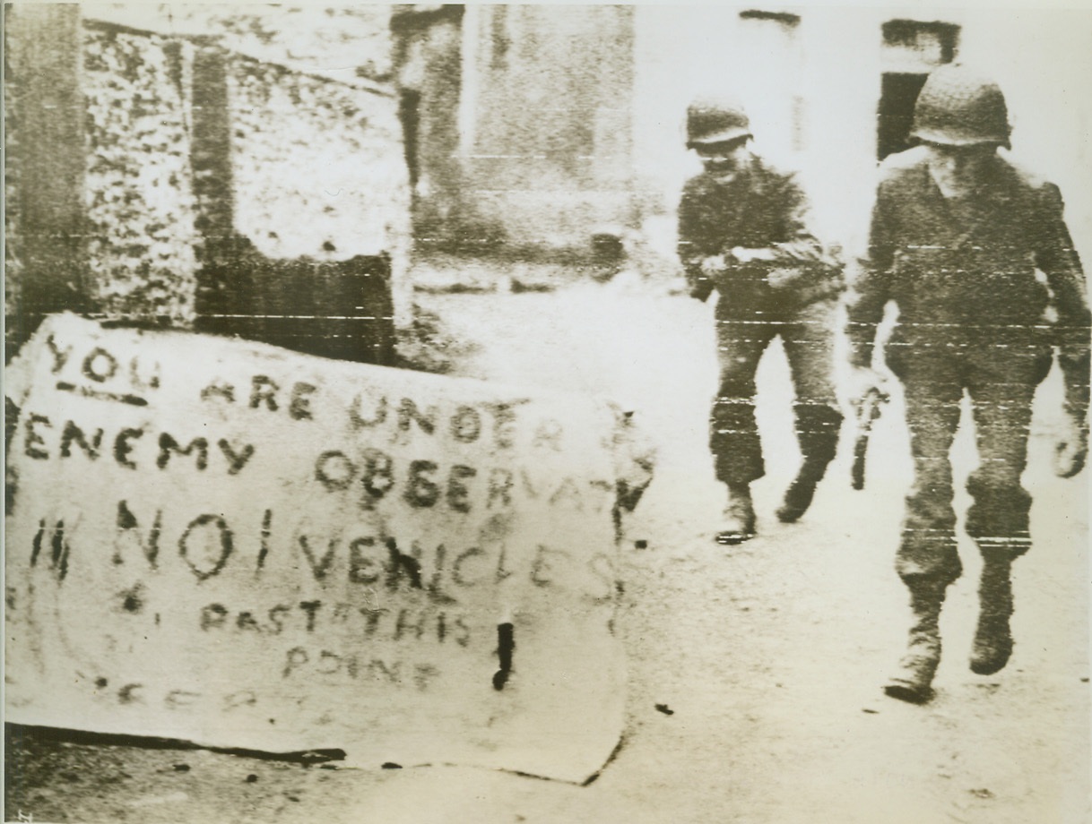 A Word To The Wise, 7/13/1944. France—Heeding the warning of this sign “Somewhere in Normandy” which reads, “You are under enemy observation—no vehicles past this point.” Two Yanks crouch low as they race past an exposed position. They are (left to right): Pfc. Russell J. Schoonmaker, of Shelton, Conn., and Pvt. James V. Pappas, Indianapolis, Ind. Credit: Army radiotelephoto from ACME;