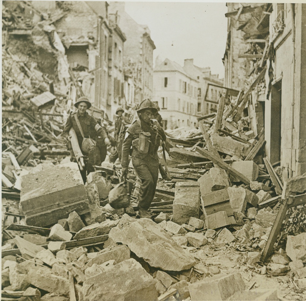 British troops make their way, 7/14/1944. Caen, France—British troops make their way precariously over the wreckage piled feet high in the streets of Caen as they advance into the town following the German retreat. They had to be constantly on the alert as the Nazis had left the streets heavily mined. Note the second Tommy carries a rolled stretcher. Credit: British official photo from ACME;