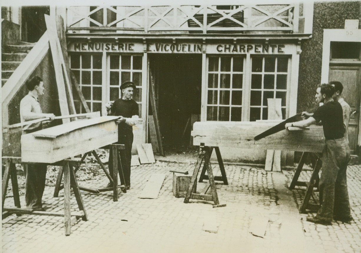 Coffins in Wholesale Lots, 7/4/1944. Grand Camp, France—Carpenters saw away at rude coffins for French civilians who were killed during the fierce fighting which followed the Allied invasion of France. So large was the number of civilian deaths that this woodmaker has enlisted the aid of three helpers to build the number of coffins needed. Credit: ACME;