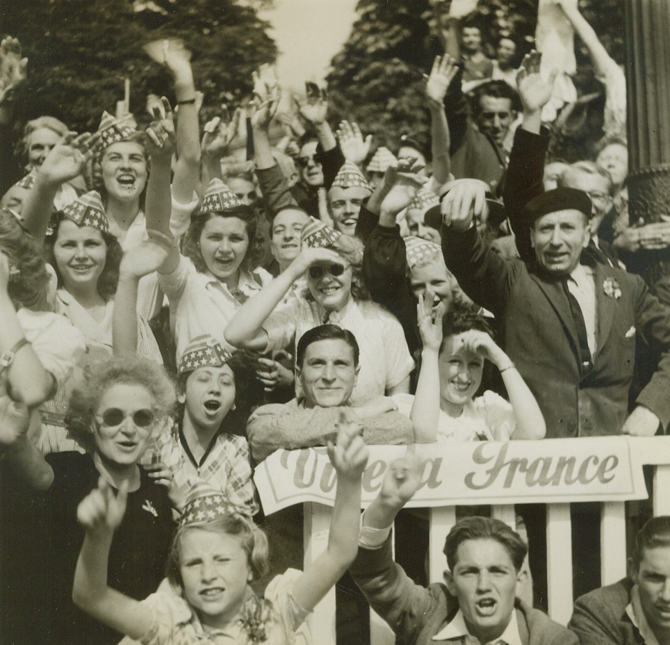 VIVE THE STARS AND STRIPES, 8/31/1944. PARIS—Wild with joy over the liberation of Paris, and singing the praises of their American allies, these celebrating Parisians let the Stars and Stripes go to their heads. Wearing the gay paper hats, they cheer and wave as the forces of liberation march past on parade. Credit: Acme photo by Bert Brandt for the War Picture Pool;