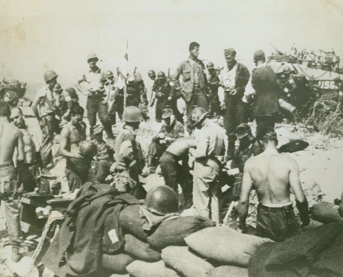 FIRST AID FOR FRIEND AND FOE, 8/16/1944. U.S. Infantrymen and German prisoners receive medical treatment from American corpsmen at a beach dressing station in Southern France, while (background), Yank troops file by on their way to consolidate the new beachhead. Credit: Acme photo by Sherman Montrose for War Picture Pool via Army radiotelephoto;