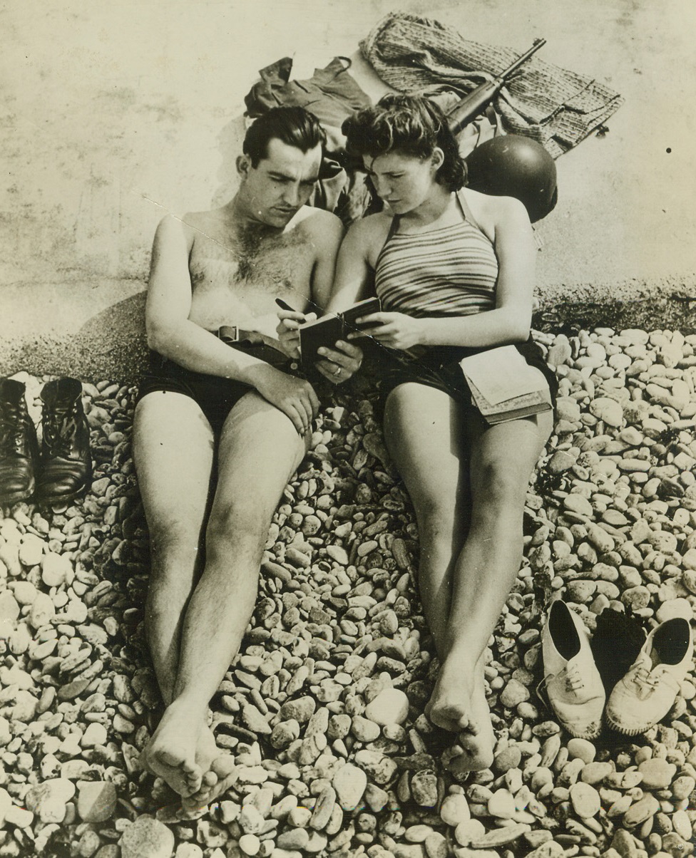 The Mamselle Gives a French Lesson, 8/12/1944. Cherbourg, France – On the rocky beach at Cherbourg, a 17-year-old French girl, Paule Marie Truffert, gives a French lesson to Sgt. Edward Watson, Terre Haute, Indiana, a member of the American forces who captured Cherbourg. Paule, who lived with her family in Cherbourg before the occupation, was sent to Valognes in May 1943 to live with her grandmother. When Yanks recaptured the port city she returned via an American jeep. Paule is a great friend of the GI’s, and now seeks a position as interpreter for the Civil Affairs Committee. She wants to start a 7-year medical course in Paris this fall. Credit: ACME;