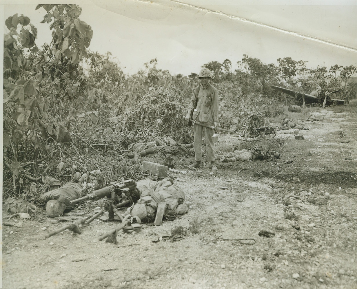 DEATH WAS THEIR SOUVENIR, 9/22/1944. PELELIU ISLAND, PALAU – Holding a Jap sword, his pockets bulging with other souvenirs of the battle for Peleliu Island, a Yank leatherneck looks at the bodies of Japs whose only souvenir was death. Two enemy machine gunners, slain in the first moments of the invasion, lie in the foreground. The Jap behind the Marine committed suicide by holding a grenade to his chest. Note wrecked plane in background. Photo by Stanley Troutman, ACME photographer for the War Picture Pool. Credit: ACME;