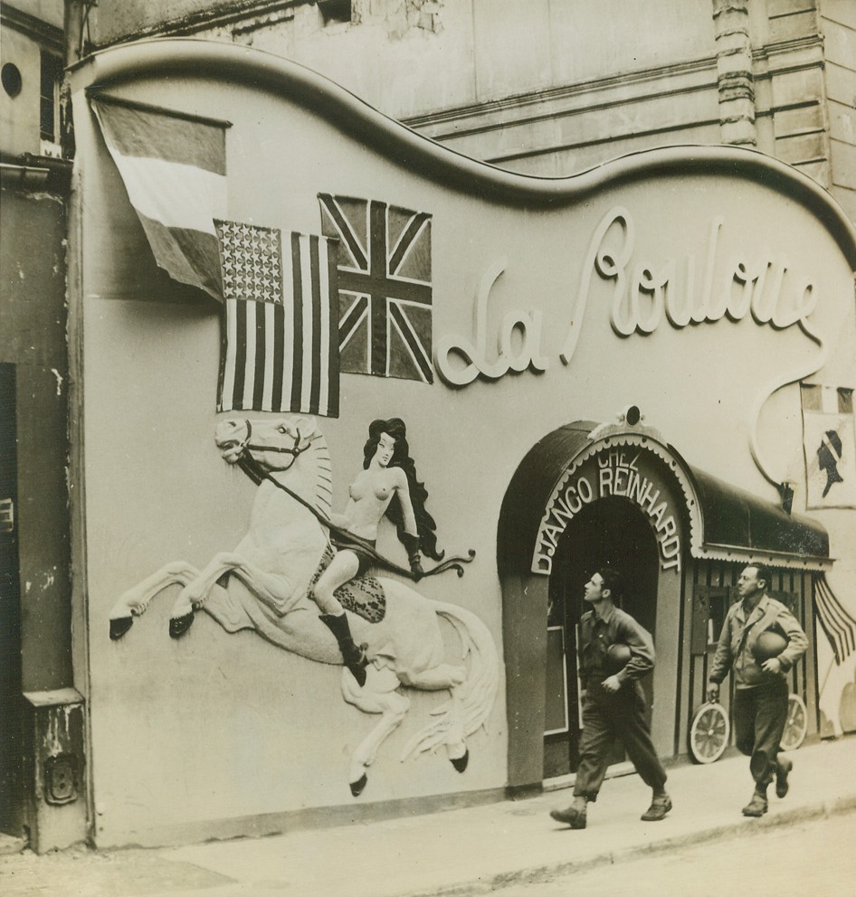 THIS IS GAY PAREE, 9/23/1944. PARIS – Unique and gaily bohemian, as only a Paris nightclub can be, the plaster front of La Roulotte is decorated with the French, American and British flags. Two GI’s strolling by observe the modern version of Lady Godiva which prances across the front. Credit: ACME;