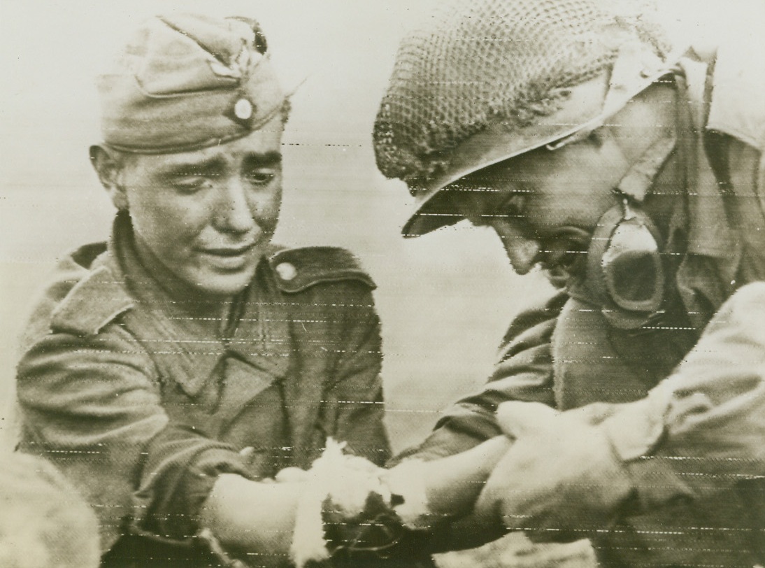 HEAL THE “CONQUERING HERO”, 9/10/1944. FRANCE—Hardly looking the part of a “conquering hero”, this youthful Nazi captive seems close to tears as a Yank examines his arm wound. The American renders first aid while awaiting the arrival of a medic.Credit: Signal corps radiotelephoto from Acme;