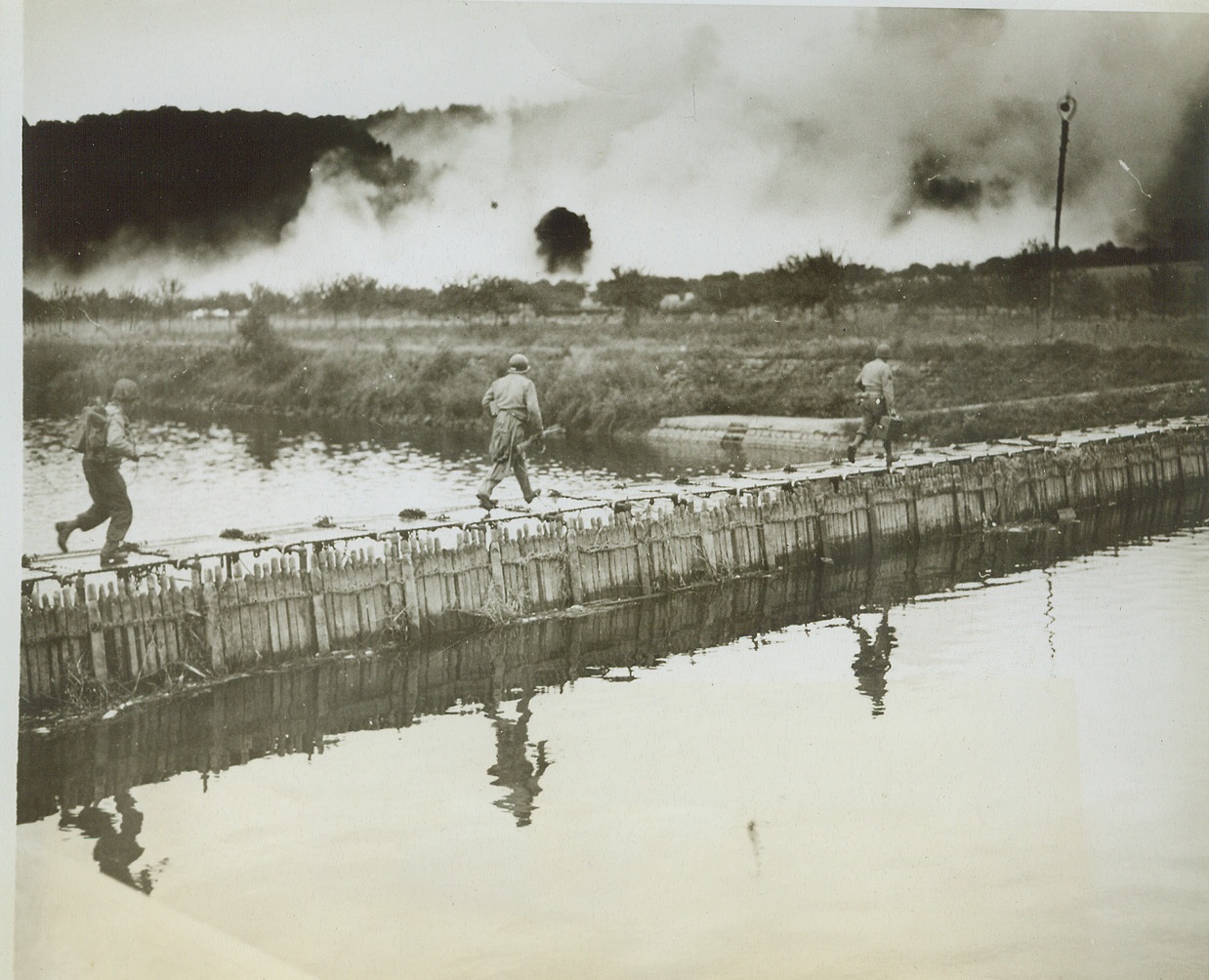 Advance in Face of Enemy Fire, 9/12/1944. Belgium – American soldiers dash across the steel plating of a bridge spanning the Meuse River in Belgium near the town of Houx as enemy shells burst around them. Nazi artillery fire falls wide of the footbridge as Yanks race across to safety. Credit (Signal Corps Photo from ACME);