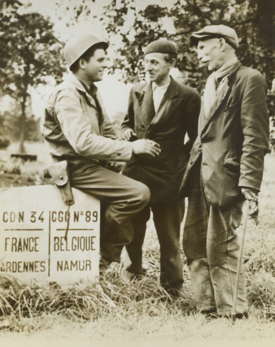 Liberation of Belgium at Hand, 9/6/1944. BELGIUM—Pvt. Gordon Conrey (left), of Milford, Vt., sits on the French-Belgium border marker and chats with two residents of Seloignes in Belgium. Swift advance across the border by Allied troops marks the beginning of the end for the Nazis in Belgium and the low countries. Credit: ACME.;