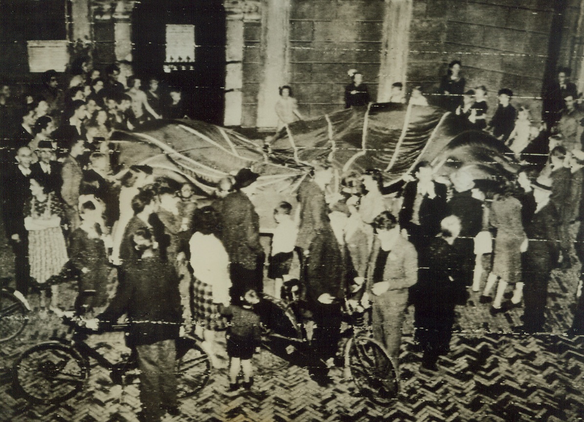 PARACHUTE DANCE, 9/24/1944. GRAVES, HOLLAND—A variation of the traditional maypole dance—heralding a far more joyous occasion than the mere advent of spring—marked the Graves celebration of its liberation by American paratroopers. Dutch women and children of the town gathered about an Allied parachute, jubilantly singing and dancing in a circle. Credit: Army radiotelephoto from Acme;