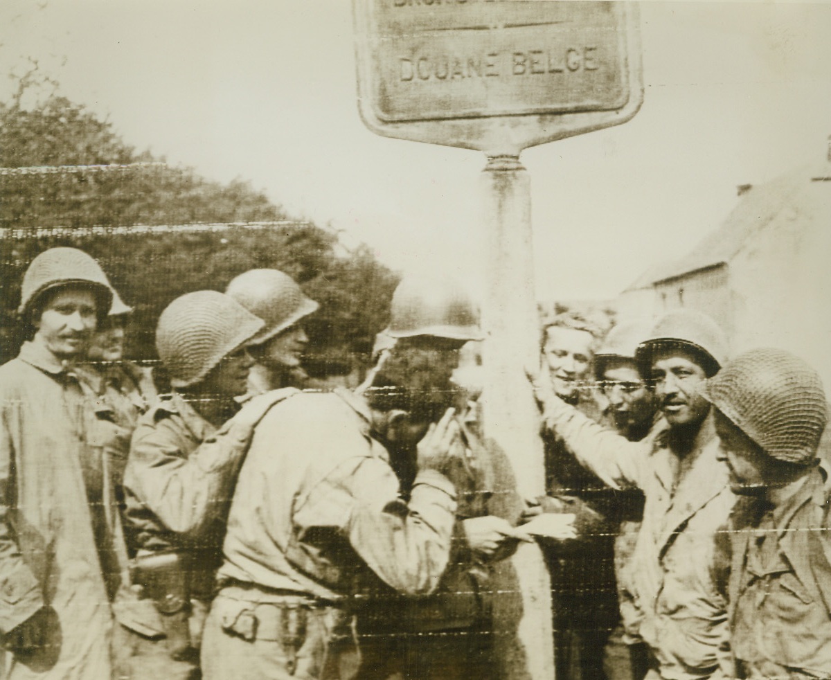 THIS IS THE WAY TO BERLIN, BOYS, 9/7/1944. BELGIUM—Hitting out in a powerful drive toward Berlin, a group of Yank soldiers pause at the Belgian border to study a map and see if they are on the right track. The sign says Belgian Customs. Credit: Signal Corps radiotelephoto from Acme;