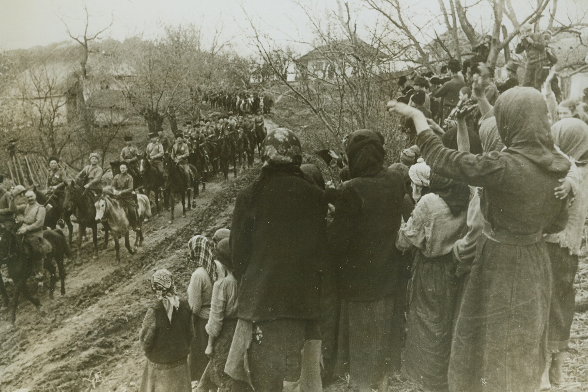 COSSACKS IN ROMANIA, 9/6/1944. ROMANIA—Residents of a tiny Romanian village, their heads covered with shawls, line a roadway to greet Don Cossack Guardsmen as they pass by. One gallant horseman (at lower right) raises his cap to the ladies. Credit: Acme;