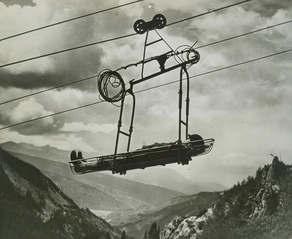 Light Aerial Tramway M-2 (3), 9/21/1944. Camp Hale, Colo.—The transportation of ammunition, food and water, and evacuation of wounded personnel, and the moving forward of artillery for mountain troops in terrain too rough for pack animals or men has long been a serious problem for mountain fighters. The army recently offered to the public its solution of this problem here when the Camp Hale test branch of the Mountain and Winter Welfare section of the Army’s Engineer Board gave a demonstration of the recently perfected Light Aerial Tramway M-2. The M-2 provides an ideal method for quickly evacuating wounded from high mountains. Shown here is a Stokes Litter, tied to the tram car and being lowered to the base terminal.  Signal corps photo from ACME.;