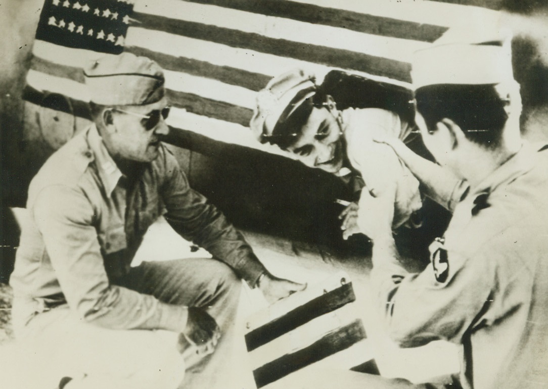 Stunt That Liberated 1000 Americans, 9/3/1944. Leaning out of the flag-covered fuselage of his plane, Lt. Gerald W. Marshall, Los Angeles, Calif., (center) demonstrates to two fellow officers of a B-24 Liberator Bomb Group of the 15th Army Air Force how Lt. Col. James A. Gunn, San Antonia, Tex., flew back from Romania to arrange for mass evacuation of over 1000 former Air Force men from a Romanian prison camp. Credit: Signal Corps Radiotelephoto from ACME;