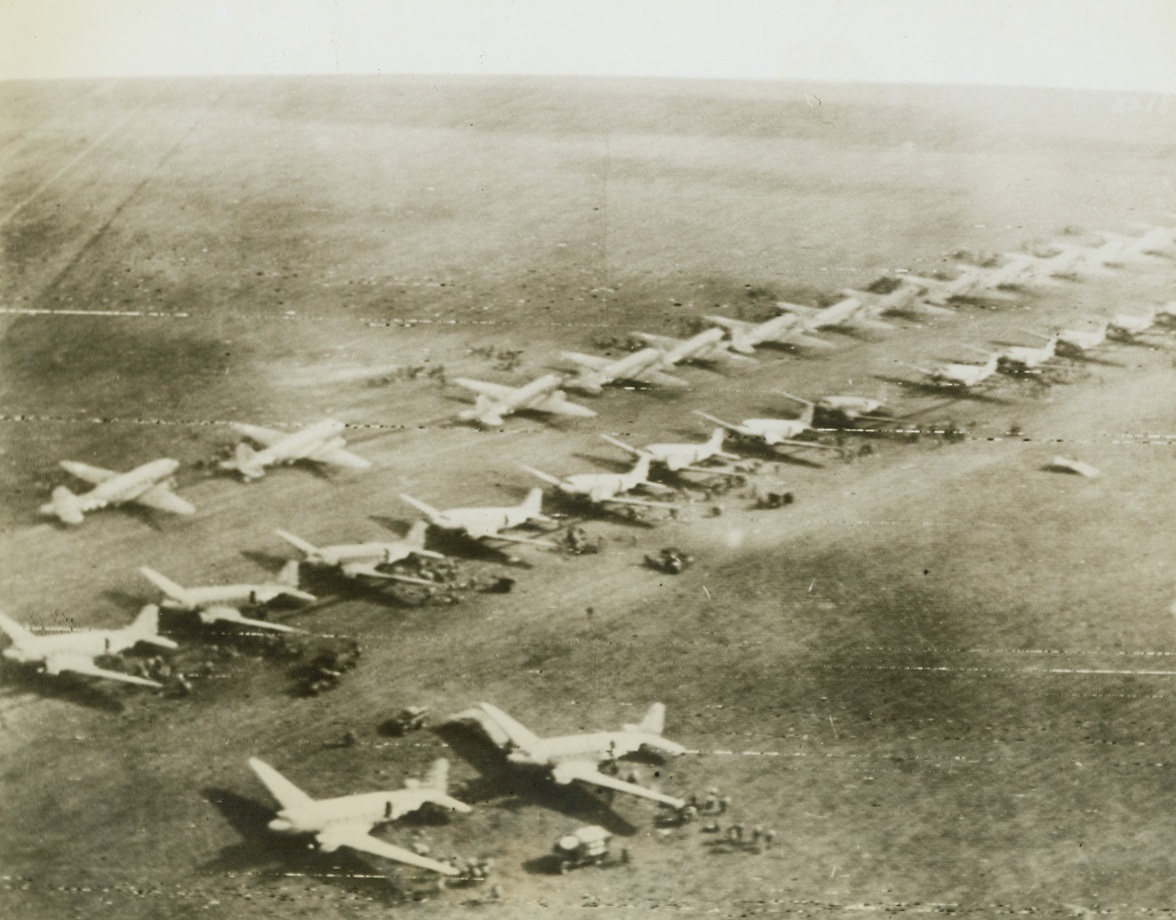 INVASION LINEUP, 9/18/1944. ENGLAND – Part of the mighty air armada that carried invading Yanks to Holland, these C-47 transports sit in orderly rows, awaiting their signal to soar.  Like tiny black dots on the airfield in England, paratroopers and ground crewmen scurry about the big warbirds as last-minute details are attended to.Credit: Signal Corps radio telephoto from Acme;