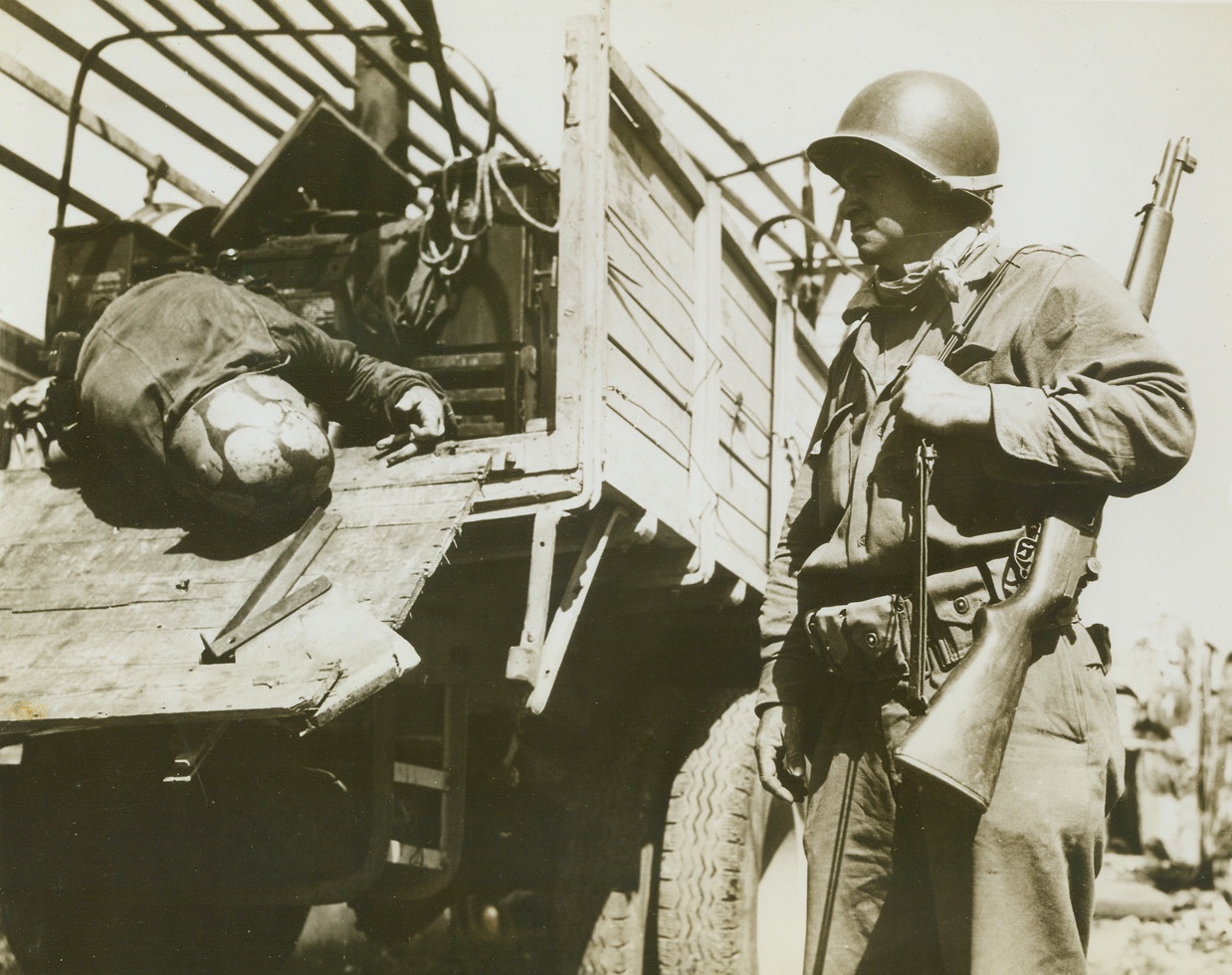 His Cargo--A Dead German, 9/12/1944. France -- Showing no emotion, Cpl. Charles S. Hoff, Cohoes, New York, looks at a dead German hanging on the lip of a truck outside of Loriol, France. Town was the site of Nazi debacle as American artillery and Air Force destroyed an entire convoy. Credit: Signal Corps photo from ACME;