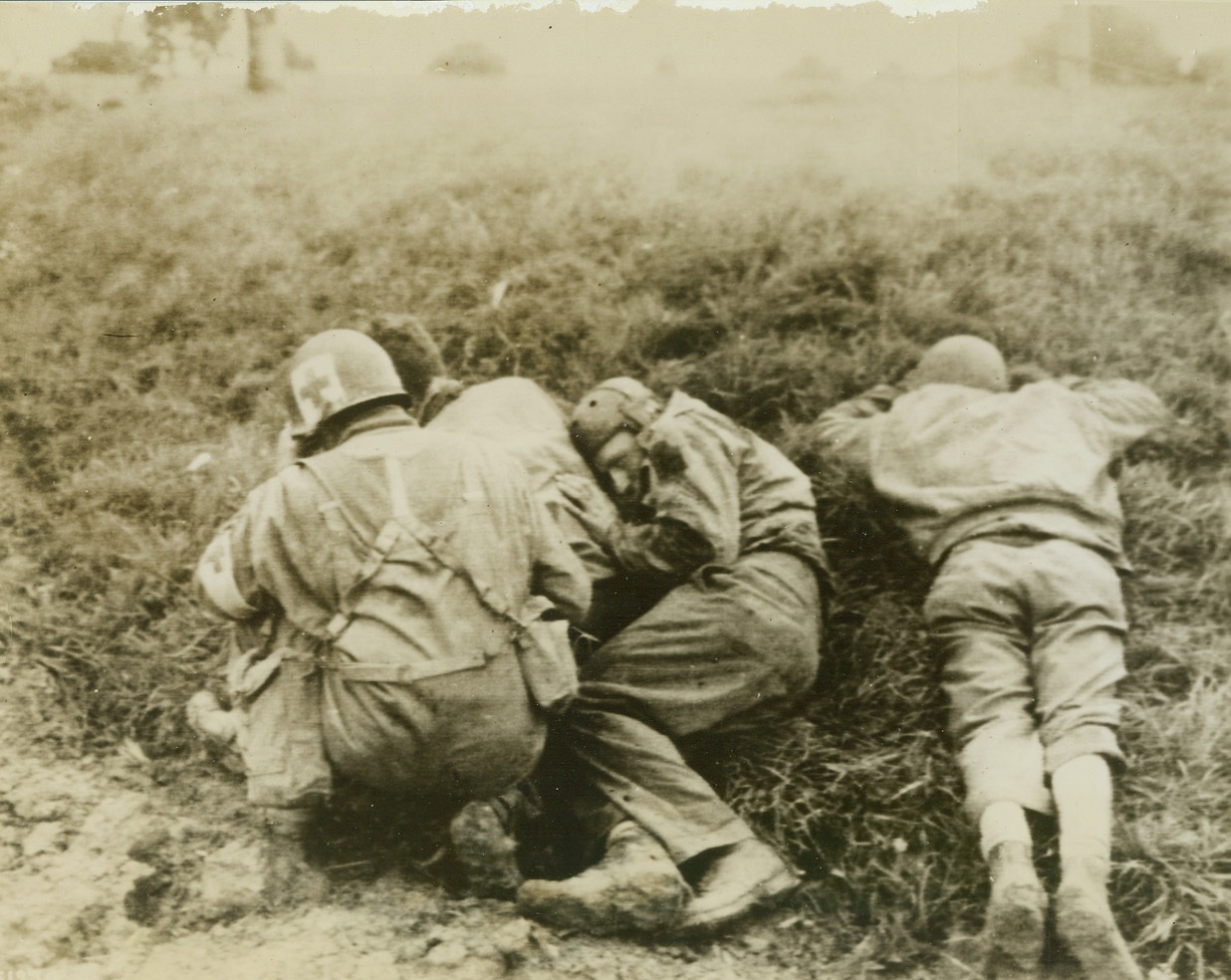Keep ‘em Low, 9/29/1944. Moncel, France – Members of a tank crew hug the ground as a German artillery barrage comes over this field near Moncel. The medic at left squats as he treats a wounded man. Credit: Army Radiotelephoto from ACME;