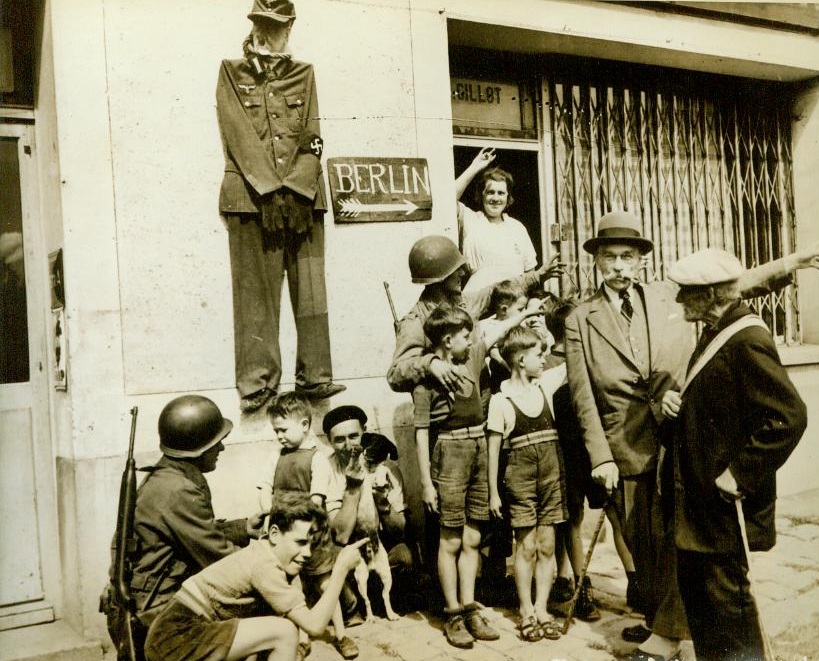 Hitler In Effigy And Road To Berlin, 9/7/1944. France -- Residents of Chateau-Thierry, France, stand under a dummy of Hitler hanging on a wall and point out the road to Berlin to two U.S. soldiers. Yanks are Pvt. Gordon Conroy (left), of Milford, N.H., and Pvt. William Rosenberg (under Berlin sign), of 10 Franklin Place, Woodmere, L.I., N.Y. 9/7/44 (ACME);