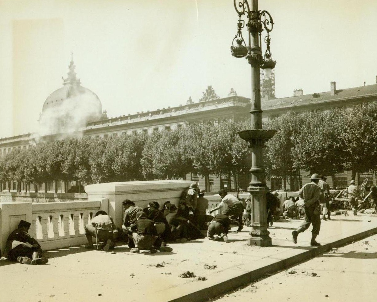 French Civilians Retaliate Sniper Fire, 9/12/1944. Lyon, France -- Before the liberation of Lyon was complete, the Maquis and French civilians had to do away with enemy snipers stationed throughout the city. Here Maquis, French troops, and civilians take cover behind a concrete wall as German sympathizers, manning two machine guns, fire on them from their stand in the Lyon hospital (background), on the south bank of The Rhone River. 9/12/44 (ACME);