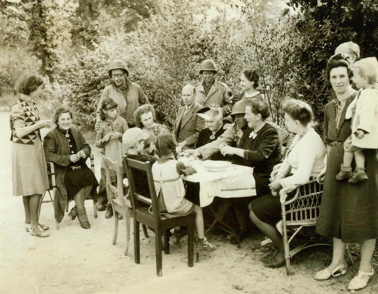 Food For The Downtrodden, 9/18/1944. Germany – Residents of a small German village, who had the courage to greet Allied warriors as liberators, look happy as they sit down to a meal of American rations. Cpl Mel White, of Harlan, Iowa, distributes the food to the hungry members of a large German family. 9/18/44 (ACME);