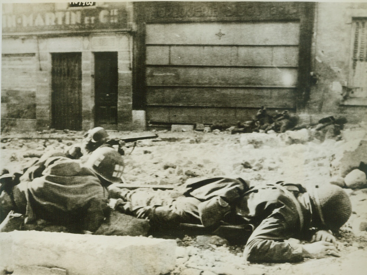 Rescue Under Fire, 9/20/1944. Madiers, France—While his buddy covers his movements, an American soldier (left) crawls into the line of fire to rescue his captain (right) who lies prone on the ground after being hit by a Nazi machine gun bullet during street fighting in Madiers, France. Bullets whizzed overhead but the Yank effected the rescue. Credit: Signal Corps radiotelephoto from ACME;