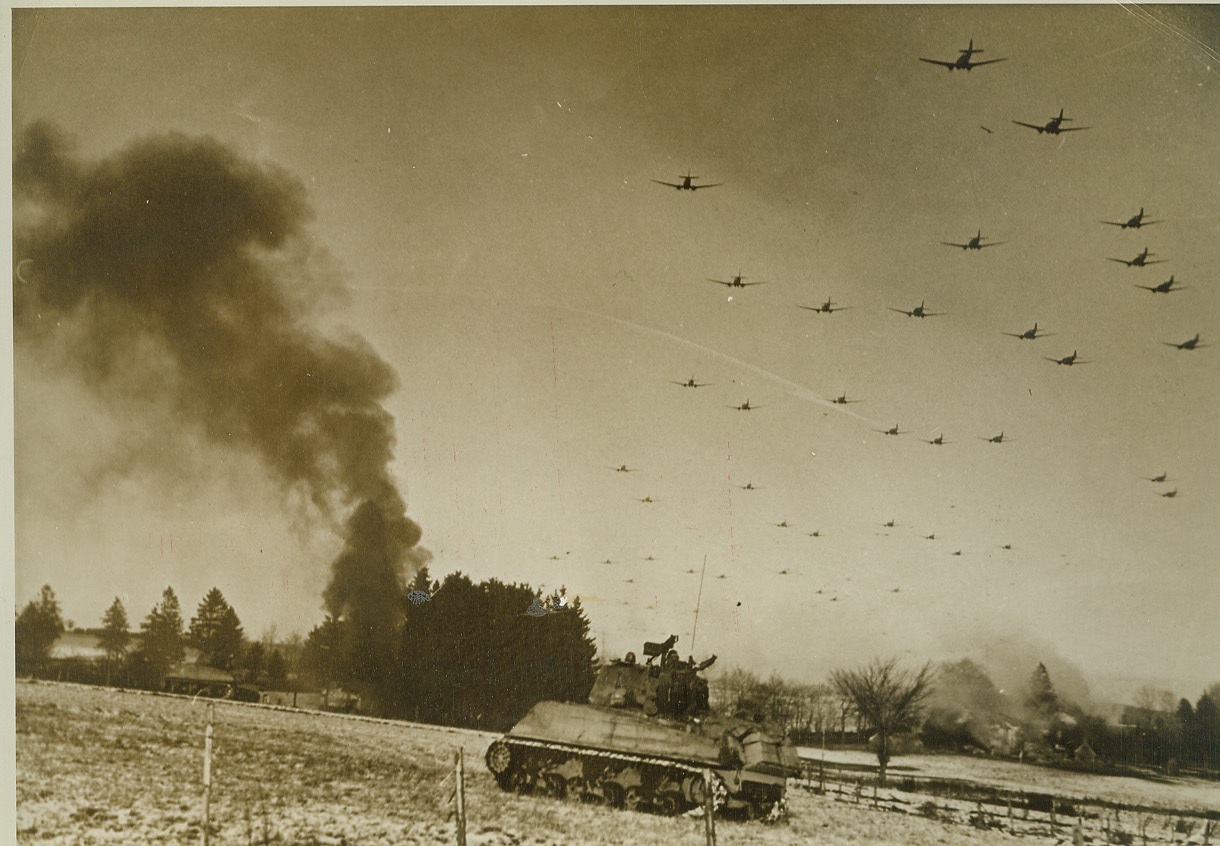 Fly Supplies to Bastogne, 1/6/1945. Belgium – An American tank rolls to attack Nazi positions as low-flying C-47 transports pass overhead carrying supplies to the hard-pressed garrison at Bastogne. Smoke from wrecked German equipment rises in the background. Credit-WP-(ACME);