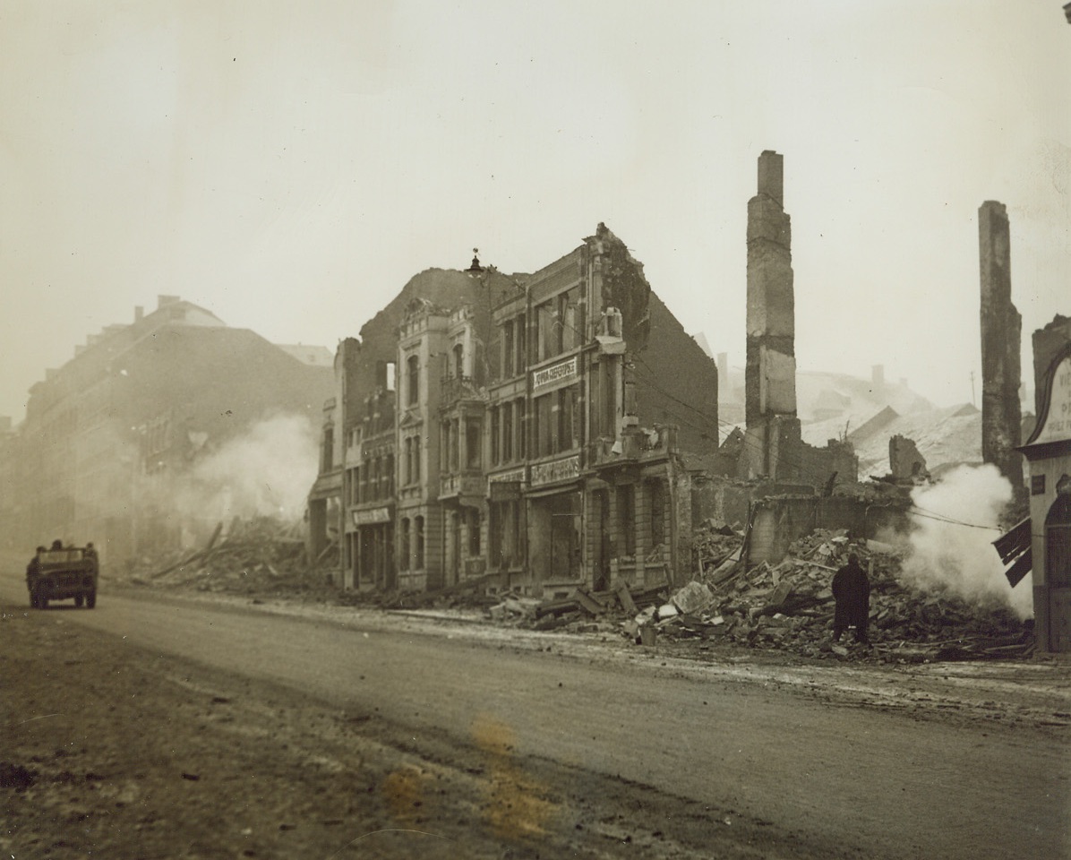 Battered Bastogne, 1/8/1945. BASTOGNE, BELGIUM—An army jeep moves down a cleared street in Bastogne after the besieged city was relieved from the pressure of German attack by men of Gen. Patton’s army. Debris is still heaped high on the sidewalks and the great number of the buildings has been shattered beyond repair by German bombs. Credit: ACME photo.;