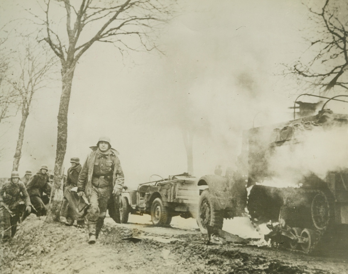 GERMANS PUSH FORWARD, 1/2/1945. WESTERN FRONT—This photo taken from a captured German film shows German infantrymen passing burning American vehicles somewhere on the Western Front during the enemy drive into American positions. Credit: Acme;