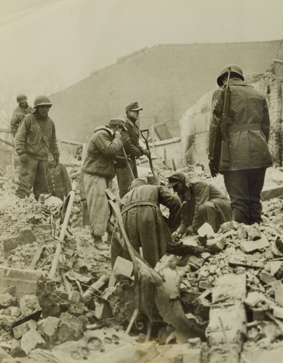 REPAIRING THE DAMAGE THEY WROUGHT, 1/8/1945. BASTOGNE, BELGIUM—Captured when Gen. Patton’s men came to relieve the besieged city of Bastogne, these German prisoners are put to work clearing away the debris wrought by their own bombs and shells. Yank soldiers oversee the job. Credit: Acme;