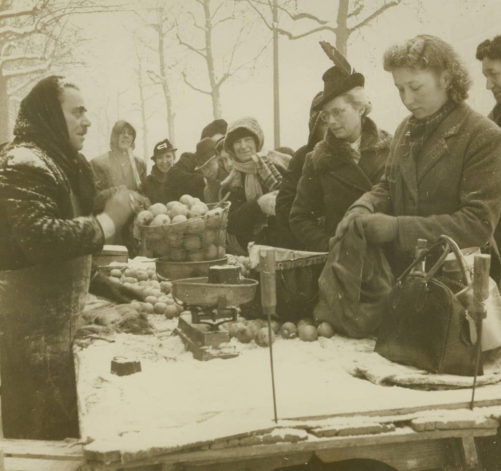 IN PARIS IT’S COLD AND FOOD IS SCARCE, 1/20/1945. PARIS—Parisians have had a tough time keeping warm this winter and almost as tough a time getting their daily ration of food. Homes, offices, style salons, all have an almost constant temperature of near zero (centigrade) and no relief is in sight. Warm clothing is scarce, coal is non-existent even in the black market and the small quantities of fire wood on hand are hard to get and prohibitive in price. As for food, French officials say the situation in Paris is “normal” after four years of occupation, but all food supplies are very limited. This series of photos by ACME photographer Sherman Montrose, for the War Picture Pool, shows what Parisians are enduring this very cold winter. New York Bureau Despite inclement weather, this apple stall in on of Paris’ largest open-air markets does a thriving business. Parisians bundled against the cold buy the few supplies on display. Credit –WP- (ACME);