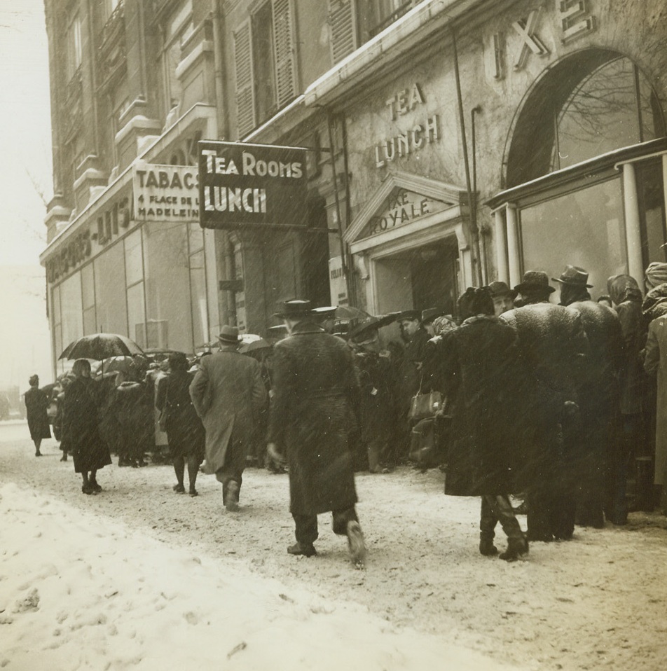 Making Do on a Cold Paris Morning, 1/20/1945. Paris, France – Weathering the first heavy snowfall of the year, Parisians line up in the morning before a café to get their ration of ersatz “coffee.” Parisians are battling limited food supplies and coal shortages during what has been a bitterly cold winter. Credit: ACME;
