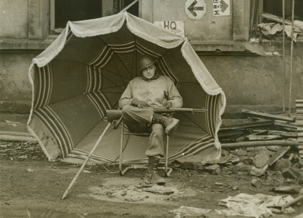 All the Comforts. . ., 3/6/1945. Grundhof, Luxembourg—Stationed in front of the railroad terminal at Grundhof, this ingenius Yank MP isn’t bothered by the rain. Snug as a bug, he’s sheltered by a large beach umbrella, found in a vacant house nearby. Photo by Charles Haacker, ACME photographer for the War Picture Pool. Credit: ACME.;