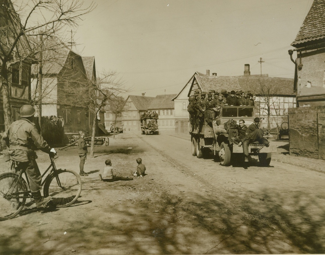 Wayside Onlookers, 4/2/1945. TREBUR, GERMANY – Two small German children look up from their game as truckloads of German prisoners, captured as the Third Army expanded its bridgehead east of the Rhine, moved through a desolate street in Trebur, Germany. Overflow of prisoners ride on the front bumpers. Photo by Charles Haacker, Acme war pool photographer.Credit (ACME) (WP);