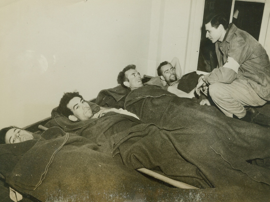 EMACIATED AMERICANS, 4/7/1945. LIMBURG AREA, GERMANY—Half-starved and badly cared for while they were held prisoners of the Germans at a camp near Limburg, these American soldiers now rest comfortably and receive good care from U.S. First Army medical men, members of the forces which captured the camp. Left to right: Pfc. Ray Hayes, Pores Knob, N.C.; Pfc. Castos Solomon, San Antonio, Tex.; Pvt. James Gore, Albany, N.Y.; and Pfc. Joseph Noughton, Luzerne, Pa. The medic is T/5 Jerome Sloven (right), Bronx, N.Y. The bed-ridden men were taken prisoners at Vetz. Credit: Acme;