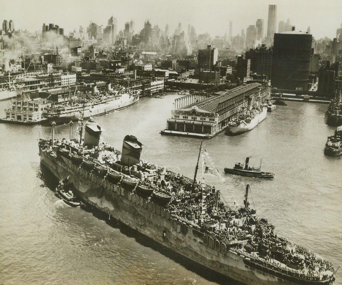 7,607 Dreams Come True, 7/11/1945. New York – Its signal flags fluttering in the breeze, and its decks lines with 7,607 eager Yanks of the 87th Division, the Navy transport West Point, largest Navy transport afloat, noses into her slip at Pier 88, North River, today (July 11). New York skyline rises in the background, a welcome sight for these battle-weary soldiers. Also aboard were a group of Japanese diplomats from the Embassy in Berlin, bound for internment. Credit: Official U.S. Navy photo from ACME;