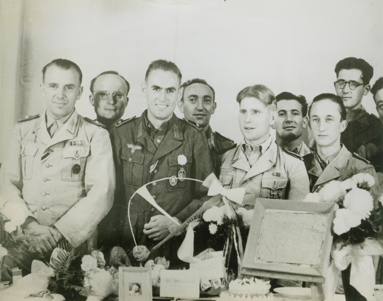 Nazi War Prisoner Married By Proxy. McAlester, Okla. - Married by proxy to his sweetheart in Germany, A prisoner of war (second from left front) in camp at McAlester, Okla., is surrounded by comrades who witnessed the ceremony. At left of bride’s picture on table is her bouquet and at right is small-tiered wedding cake. Oil painting, a wedding present, has been censored. Bridegroom wears a ribbon indicating he has received the Iron Cross, second class; the medals are a wound medal and an athletic medal. Credit: ACME;