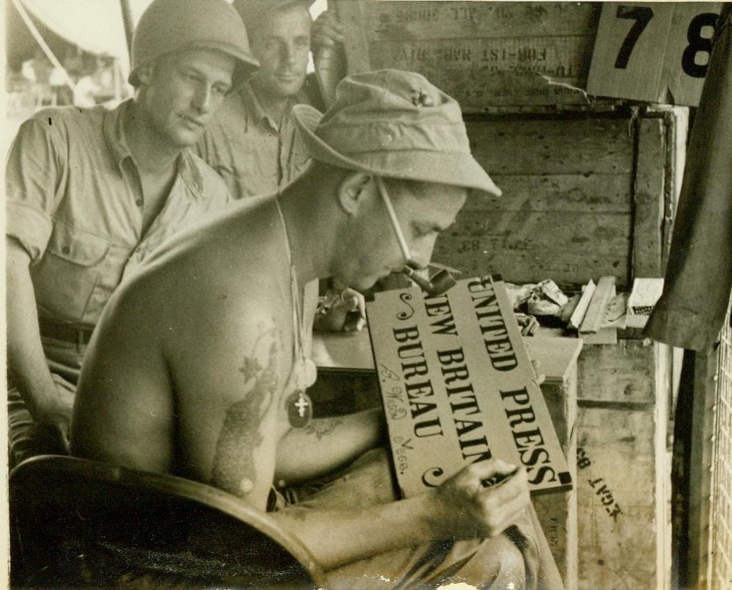 Gag sign. SOMEWHERE IN NEW GUINEA – Just before he left New Guinea to cover the Marine Corps invasion of New Britain, United Press Correspondent Bill Wislen (background, left) had a leatherneck friend paint this gag sign for him. Corporal Walter Klaptosky of Scranton, Pa., puts the finishing touches to the sign, which reads: “United Press – New Britain Bureau”.;