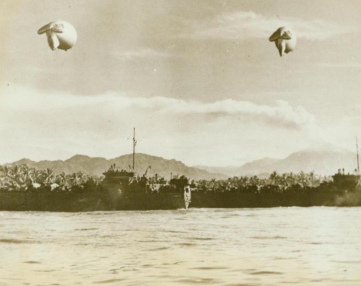 LST's Guardian Angels. Bougainville: Two barrage balloons dangle lazily in the air over several LST's unloading supplies for American forces, who have been pushing the Japs backwards into the jungle from the beachhead at Bougainville. The balloons keep Jap planes from making low-level air attacks. Far in the distance can be seen the smoking crater of Bagano, an active volcano on this spot. (ACME);