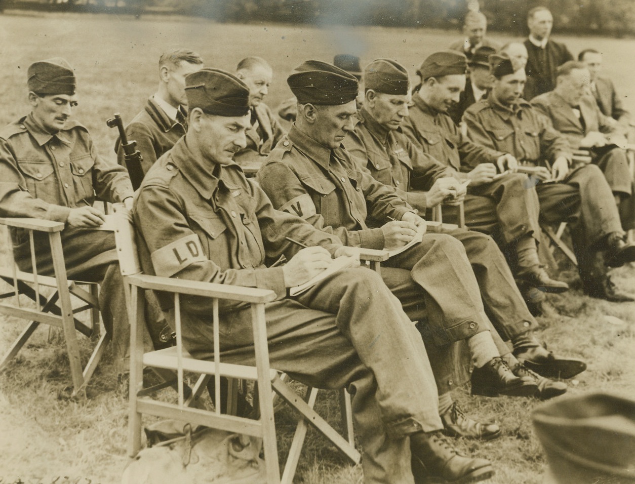 Britain’s Home Guard Studies Guerrilla Warfare, 8/7/1940  Middlesex, England—Members of Britain’s ever-increasing Home Guard who desire to have instruction in guerilla warfare tactics are attending a privately run school at Osterley Park. The courses are designed to show how to improvise to meet new situations and to use guile in overcoming the enemy. All instructors are experienced veterans. Here, some of the students take notes during an open air lecture. Credit: ACME;