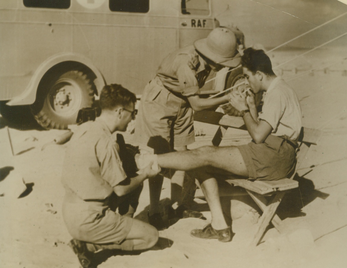 MAKING THE ENEMY AT HOME, 12/27/1940  THE WESTERN DESERT—An Italian airman shot down in a battle over Mersa Matruh Oct. 31 receives a bandage for his wound and a cigarette for his nerves from attendants at an R.A.F. desert medical station. According to British-censored caption accompanying this photo twelve Italian planes were shot down in the engagement, eight of them in flames, and this airman was the only survivor of the 35 flying the planes. Credit: Acme;